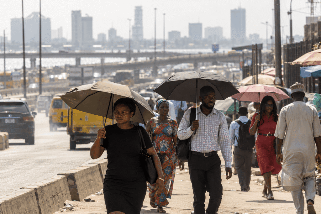 Why Is My Face So Oily by Afternoon? Causes in Hot, Humid Weather 2 Pedestrians with oily skin walk along a sunny city sidewalk, some carrying umbrellas for shade against the humid weather, with a blurred highway and skyline in the background.